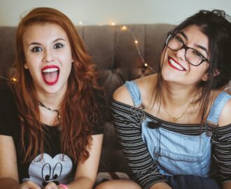 two female teenagers sitting on couch with big smiles as they use tips to save money SheeksFreaks Financial Skills for Young Adults