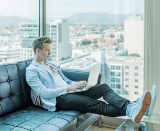 young man sits on couch in highrise building with great views works on his laptop checking his financials SheeksFreaks Financial Skills for Young Adults